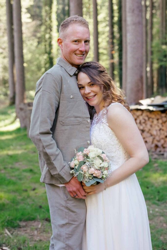 Hochzeitspaar im Wald, lachend, mit Brautstrauß, natürliche Atmosphäre.