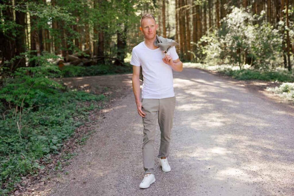 Mann im weißen T-Shirt und grauen Hosen im Wald mit Vogel auf Schulter.