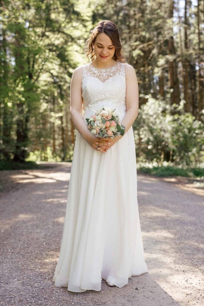 Braut im weißen Hochzeitskleid hält Blumenstrauß im Wald, natürliche Hochzeitsaufnahme.