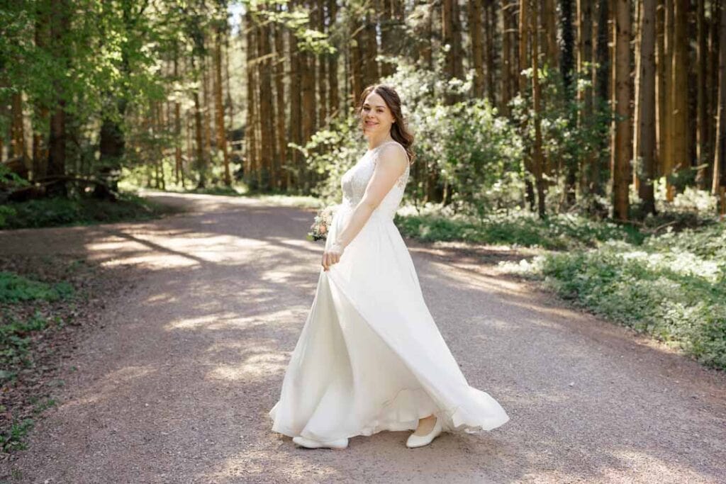 Braut im weißen Kleid auf Waldweg bei Sonnenlicht, natürliche Hochzeitsaufnahme.