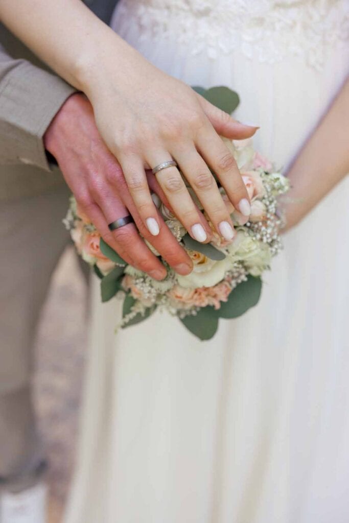 Nahaufnahme der Hände eines Brautpaares mit Eheringen und Blumenstrauß bei der Hochzeit.