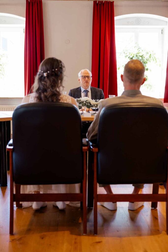 Brautpaar bei standesamtlicher Trauung, sitzend vor Standesbeamtem, mit roten Vorhängen im Hintergru.
