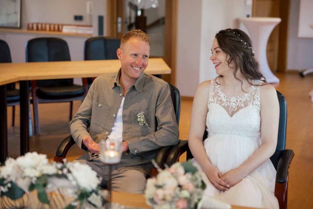 Hochzeitspaar sitzt bei einer Zeremonie, lächelnd und glücklich, in einem festlichen Raum.