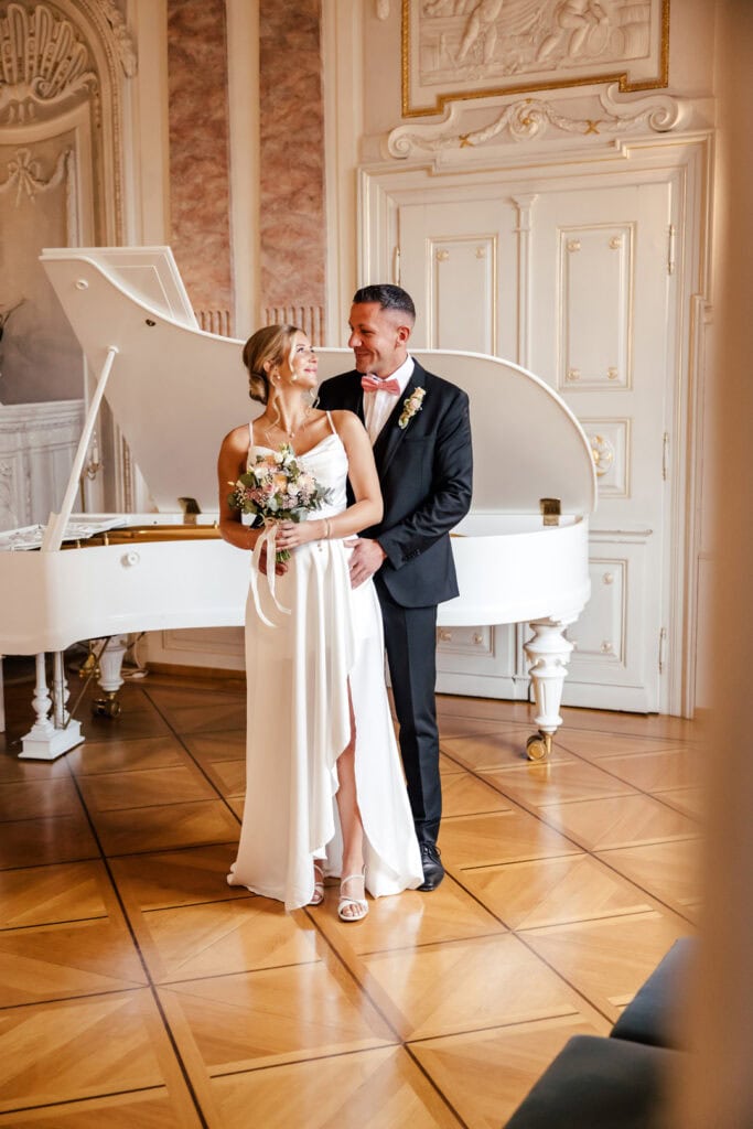 Hochzeitspaar vor weißer Klavier im prunkvollen Saal.