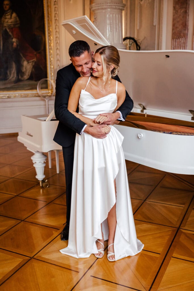 Hochzeitspaar umarmt sich vor weißem Klavier in eleganten Räumen.