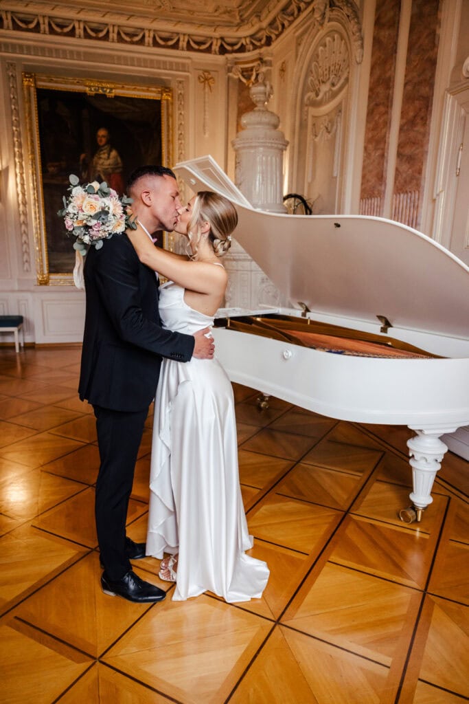 Braut und Bräutigam küssen sich vor weißer Klavier im eleganten Saal.