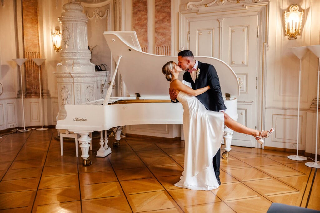 Hochzeitspaar küsst sich vor weißem Flügel in eleganter Location.