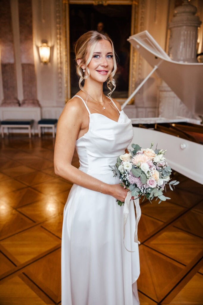 Braut in weißem Kleid mit Blumenstrauß vor weißem Klavier in festlichem Saal.