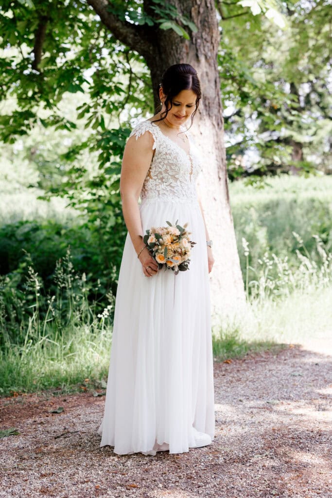 Braut in weißem Hochzeitskleid mit Blumenstrauß im Grünen.