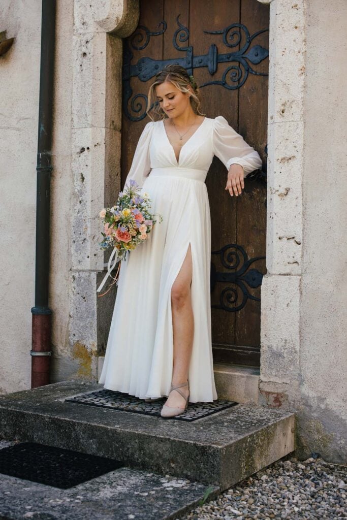 Braut in weißem Kleid mit Blumenstrauß vor historischer Tür.